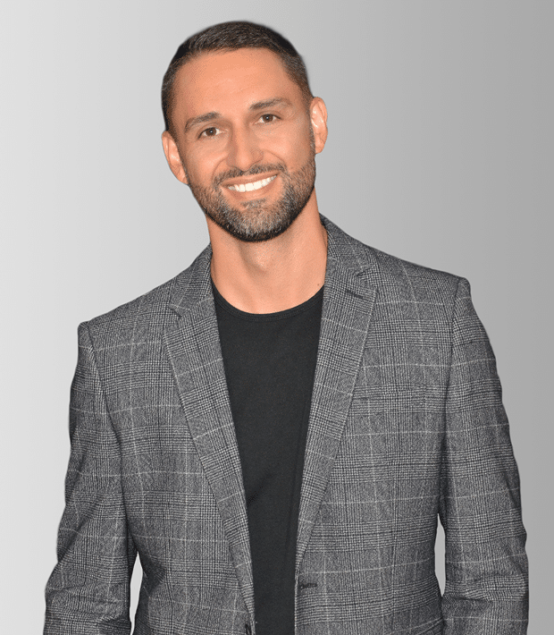 A smiling man with a short beard and dark hair wears a gray plaid blazer over a black t-shirt.
