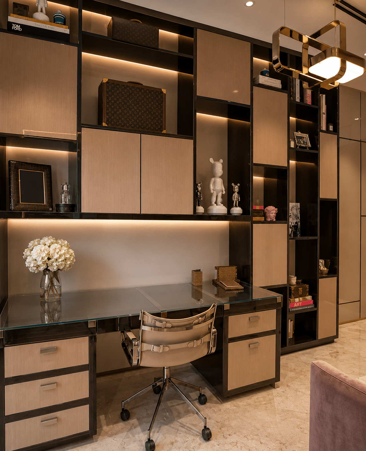 A modern home office features a desk with a glass top, a beige rolling chair, and a large built-in shelf unit with various decorative objects.